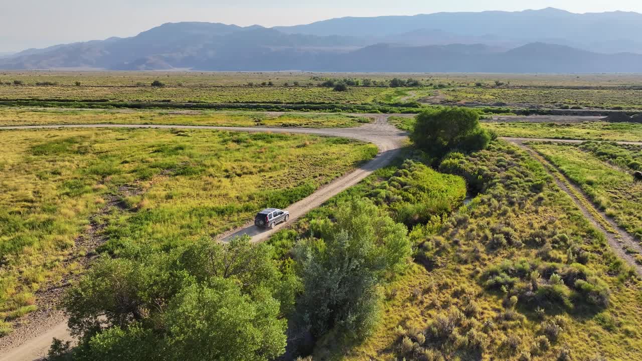 Drone pushes in and tracks a car driving along a dirt road, a small creek, and bridge on the Owens Valley floor near Bishop California. Warm late day light open rangeland with distant Sierra peaks