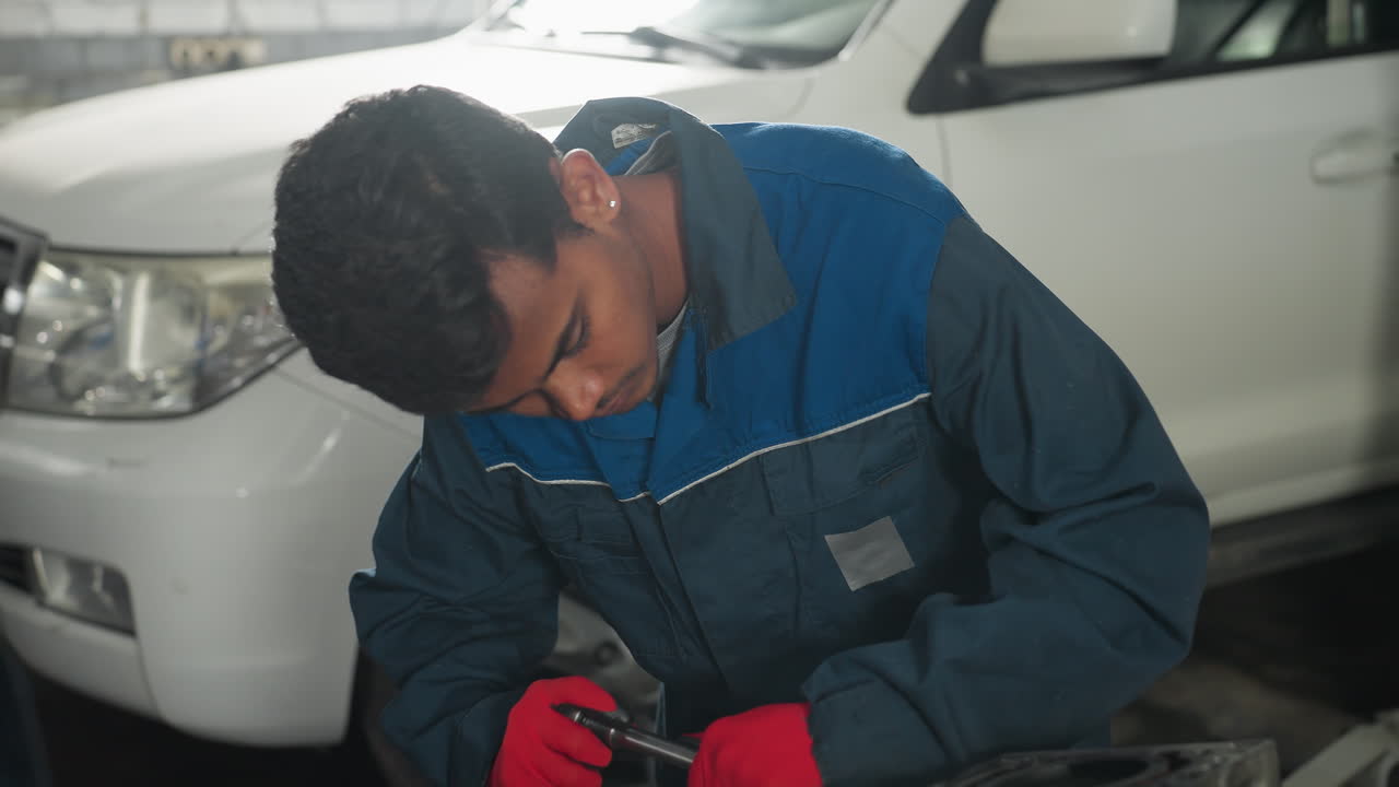 ingeniero automotriz en uniforme azul y guantes rojos centrado en la reparación del motor del coche mientras su colega mira desde atrás, el fondo presenta un vehículo estacionado en un taller automotriz profesional