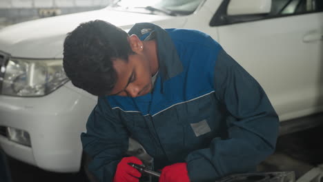 automotive engineer in blue uniform and red gloves focused on repairing car engine while colleague watches from behind, background features parked vehicle in a professional automotive workshop