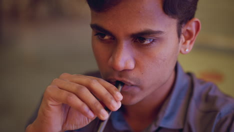 young man in indoor setting holding straw near lips, looking off to side with focused expression, wearing collared shirt and small earring, seated at table, calm atmosphere, soft lighting in background