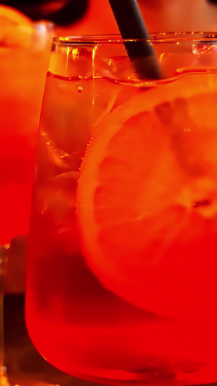 Una bevanda fredda con cubetti di ghiaccio e una fetta d'arancia brillante è mostrata con una cannuccia nera, illuminata da una calda luce arancione che crea un'atmosfera accogliente. Video verticale, ideale per i social media