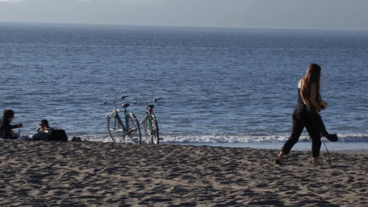 A young woman walking on the beach with water and various people in the background.