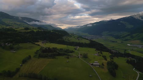 Aerial-drone-along-a-mountain-valley-in-the-Austrian-alps-with-fields,-trees-and-scenic-dramatic-sunset-clouds-in-nature-at-a-skiing-sport-hiking-and-trekking-vacation-spot-on-a-sunny-lush-summer-day