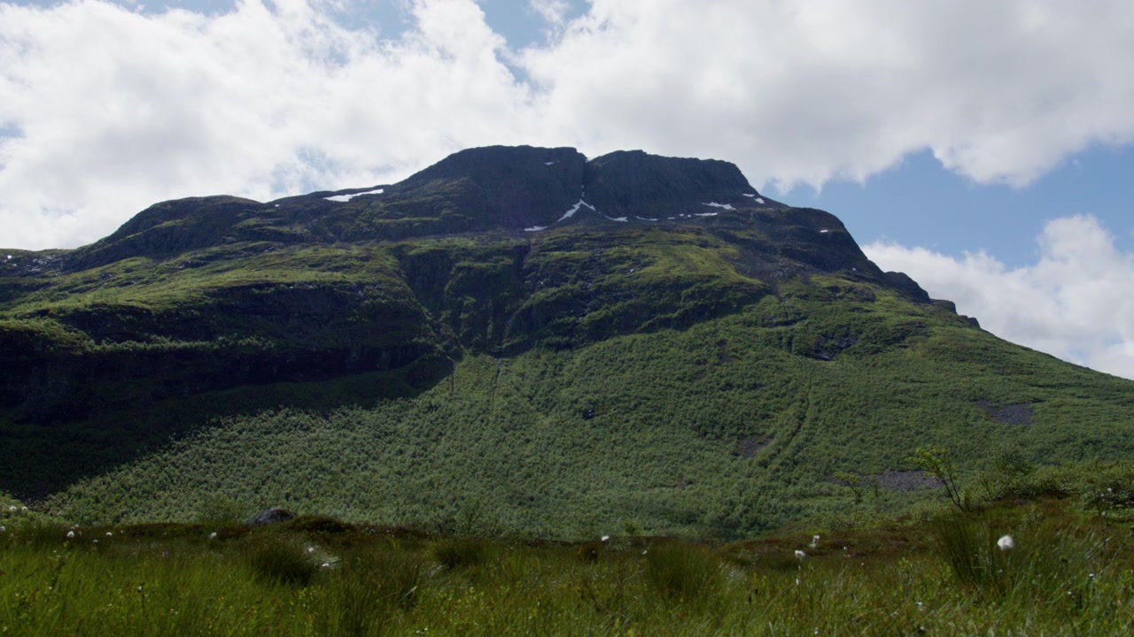 Encostas íngremes e verdes das montanhas em Sunnmøre elevam-se acima de um primeiro plano gramado sob nuvens esparsas de verão na Noruega.