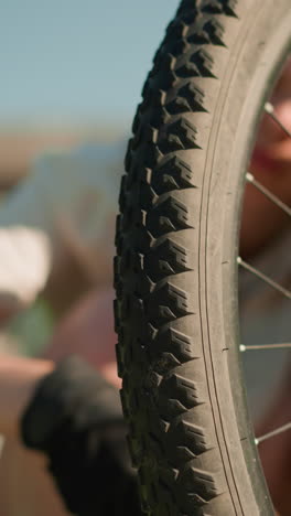 primer plano de un neumático trasero de bicicleta con un patrón de banda de rodadura robusto inspeccionado para la presión del aire por una persona que lleva guantes negros, los dedos presionando firmemente la pared lateral del neumático, un edificio suavemente borroso en el fondo