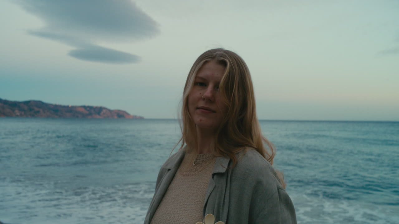 Ritratto al rallentatore di una giovane donna pensierosa che si volta verso la macchina fotografica in riva al mare al tramonto, con le onde che si infrangono dolcemente dietro di lei