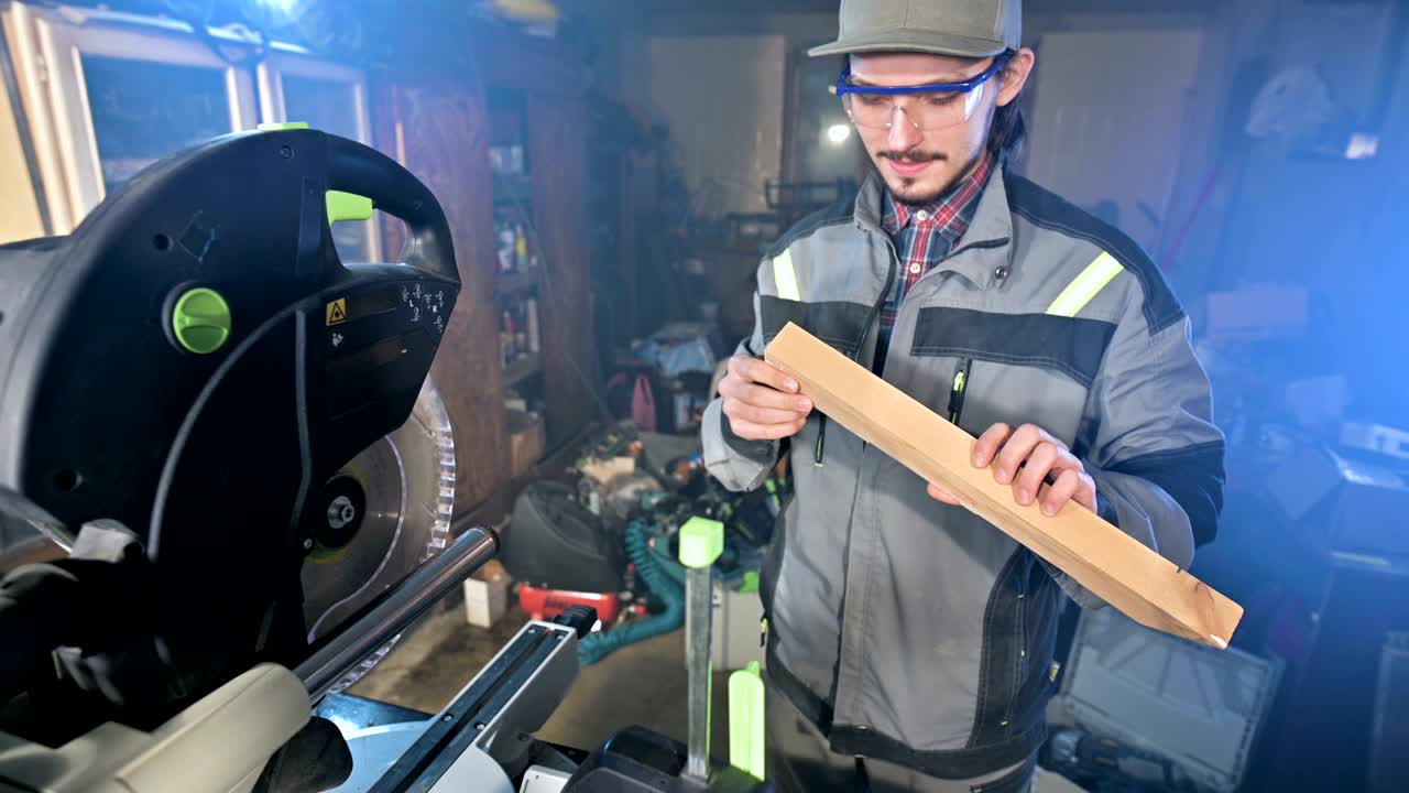 Concentrated young bearded joiner in goggles and a cap checks the work piece