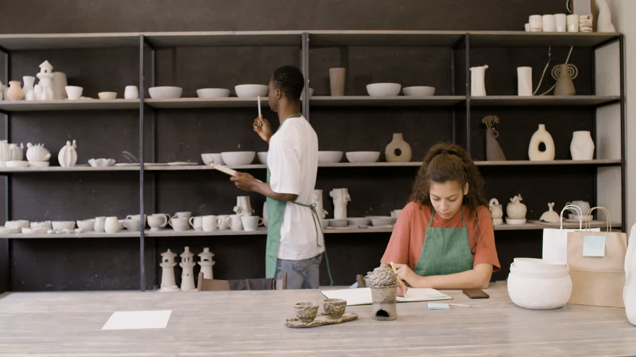 여자가 테이블에 앉아 공책에 글을 쓰고 도자기 가게에서 전화를 사용하는 동안 젊은 미국 남성 점원이 도자기 목록을 만들고 있습니다.