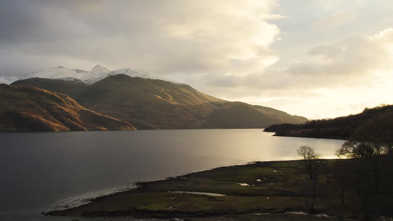 눈 덮인 산, 나무, 천연림이 물에 반사되는 황금빛 일몰과 함께 겨울철 스코틀랜드 고원의 로크 에티브와 글렌 에티브 위로 공중 드론 영상이 떠오릅니다.