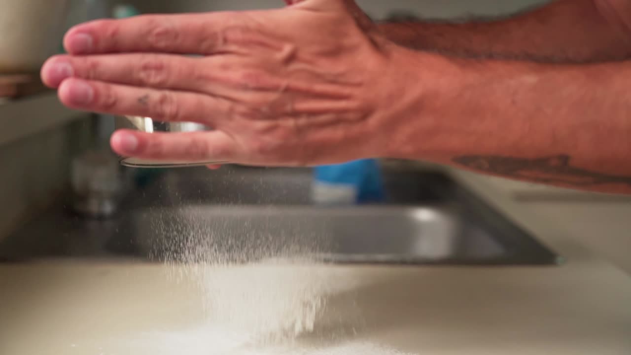 Slowmotion af kaukasisk hånd, der sigter hvidt mel med en si i et køkken.