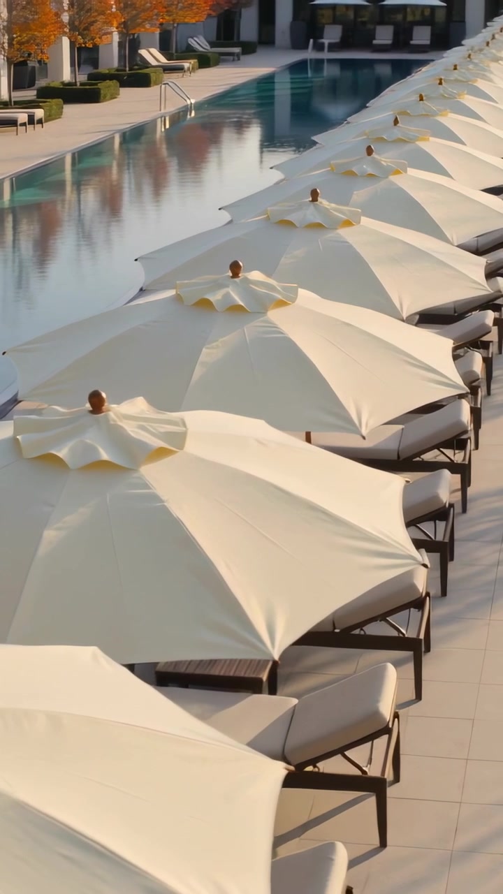 Vertical video: Camera pulling back revealing cream umbrellas by pool, showing seats, reflections