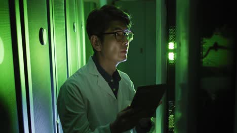 Asian-male-it-technician-in-lab-coat-using-tablet-checking-computer-server