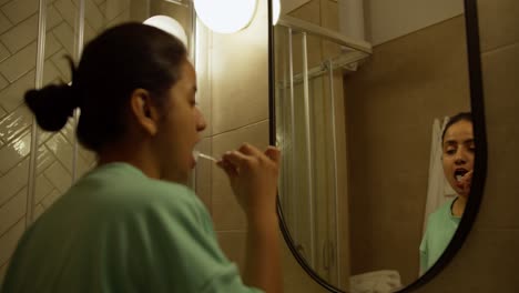 Happy-brunette-girl-with-Brown-skin-in-a-green-jacket-brushes-her-teeth-during-her-morning-routine-in-the-morning-in-the-bathroom-with-a-mirror