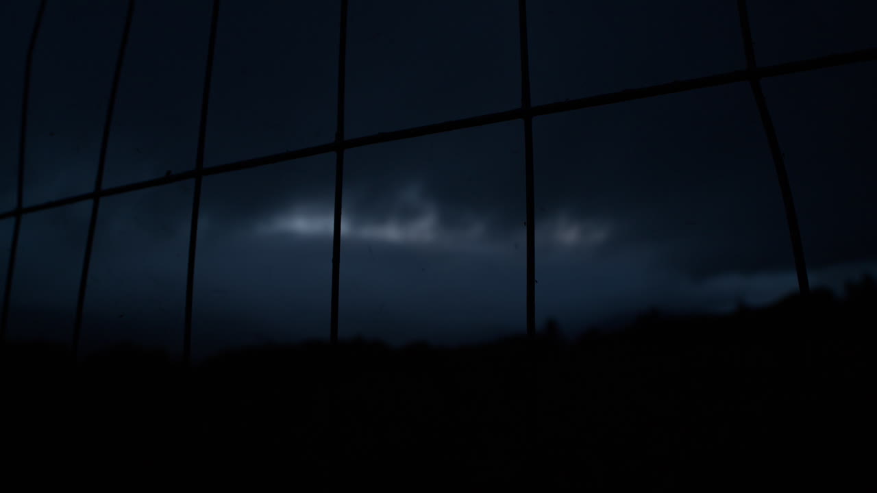 Looking through metal fence into dark night sky, moving clouds during nightfall, dark silhouette