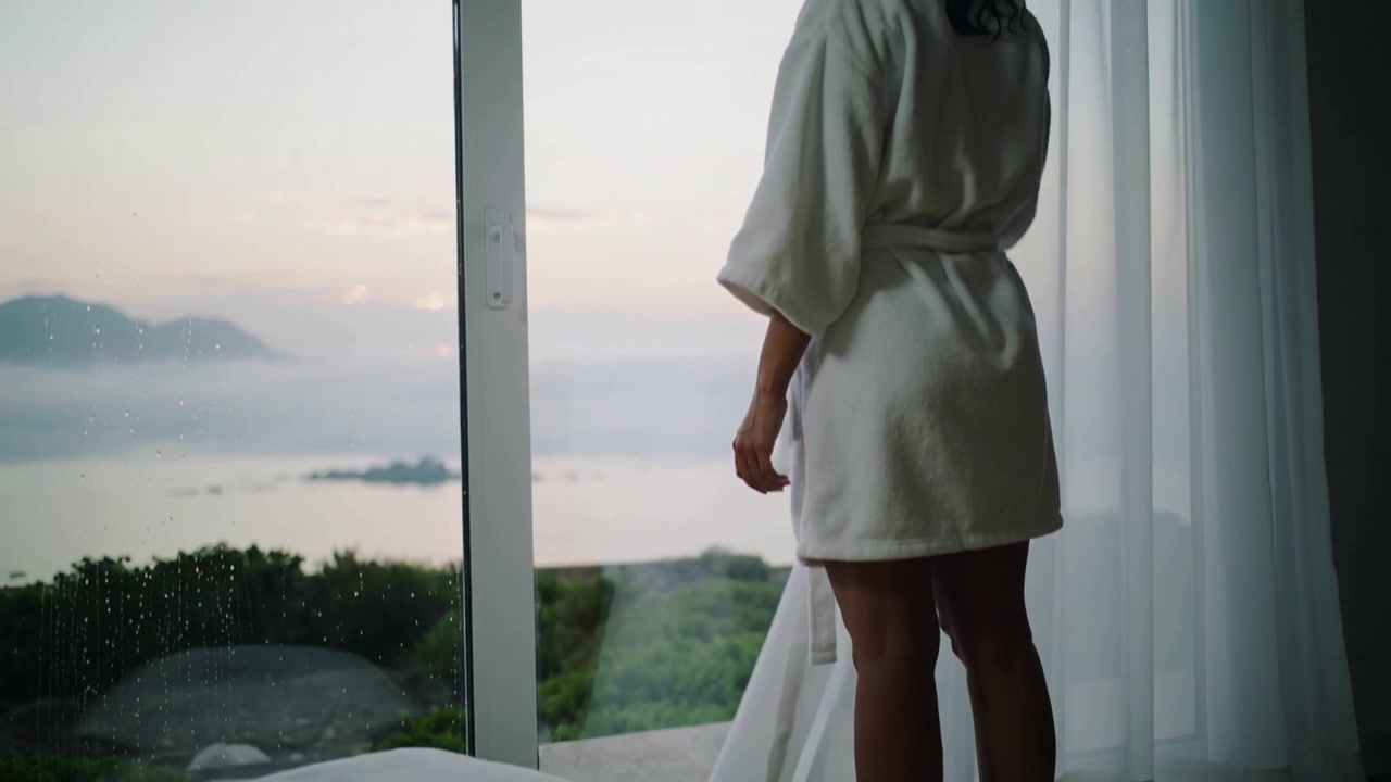 Noticing raindrops woman in white robe shifting toward sliding glass door, seeking ocean