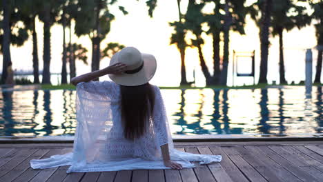 Mujer-Solitaria-Y-Elegante-Con-Sombrero-De-Verano-Y-Capa-Sentada-Junto-A-La-Piscina-De-Un-Hotel-De-Lujo,-Tiro-Trasero-Estático