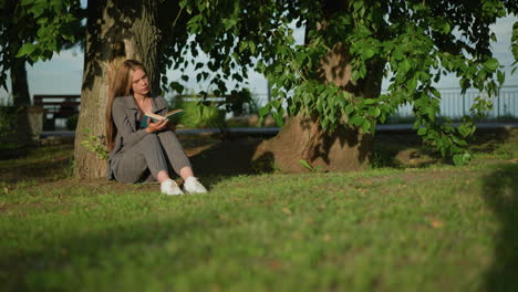 lady sitting outdoors, head slightly tilted, leaning against tree on grassy field, reading book, tree leaves sway gently in breeze, background features greenery and slightly blurred iron rail