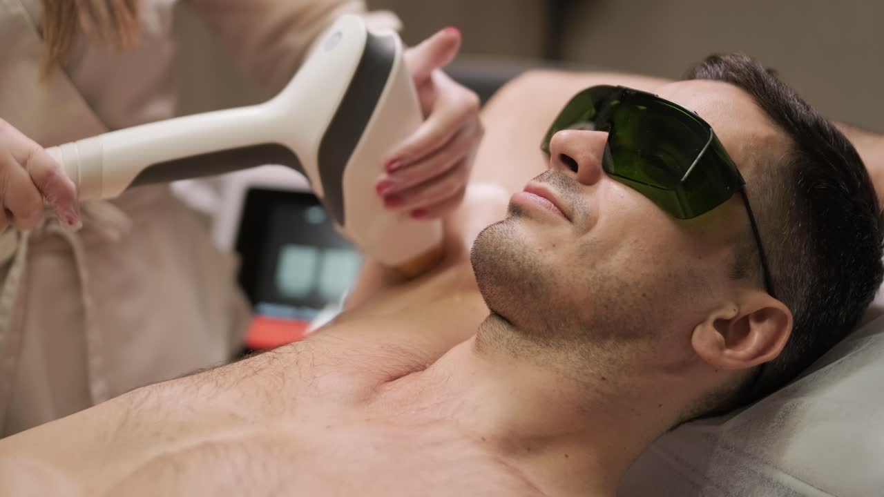 전문 레이저 기술자가 남성 클라이언트의 팔꿈치 부위에 안전 장비를 착용한 임상 환경에서 탈모 치료를 수행합니다.