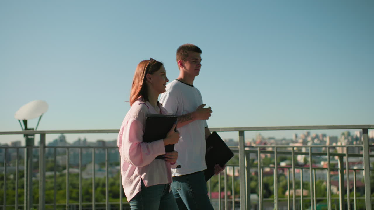 젊은 성인들은 철의 레일링 근처에서 걸어가고, 남자는 웃는 여자와 따뜻하게 대화하고, 노트북을 들고, 밝은 파란 하늘 아래의 초록색과 도시 풍경이 배경입니다.