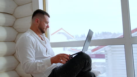 focused bearded caucasian male sits at the window. man works on the laptop and sometimes glances to the window.