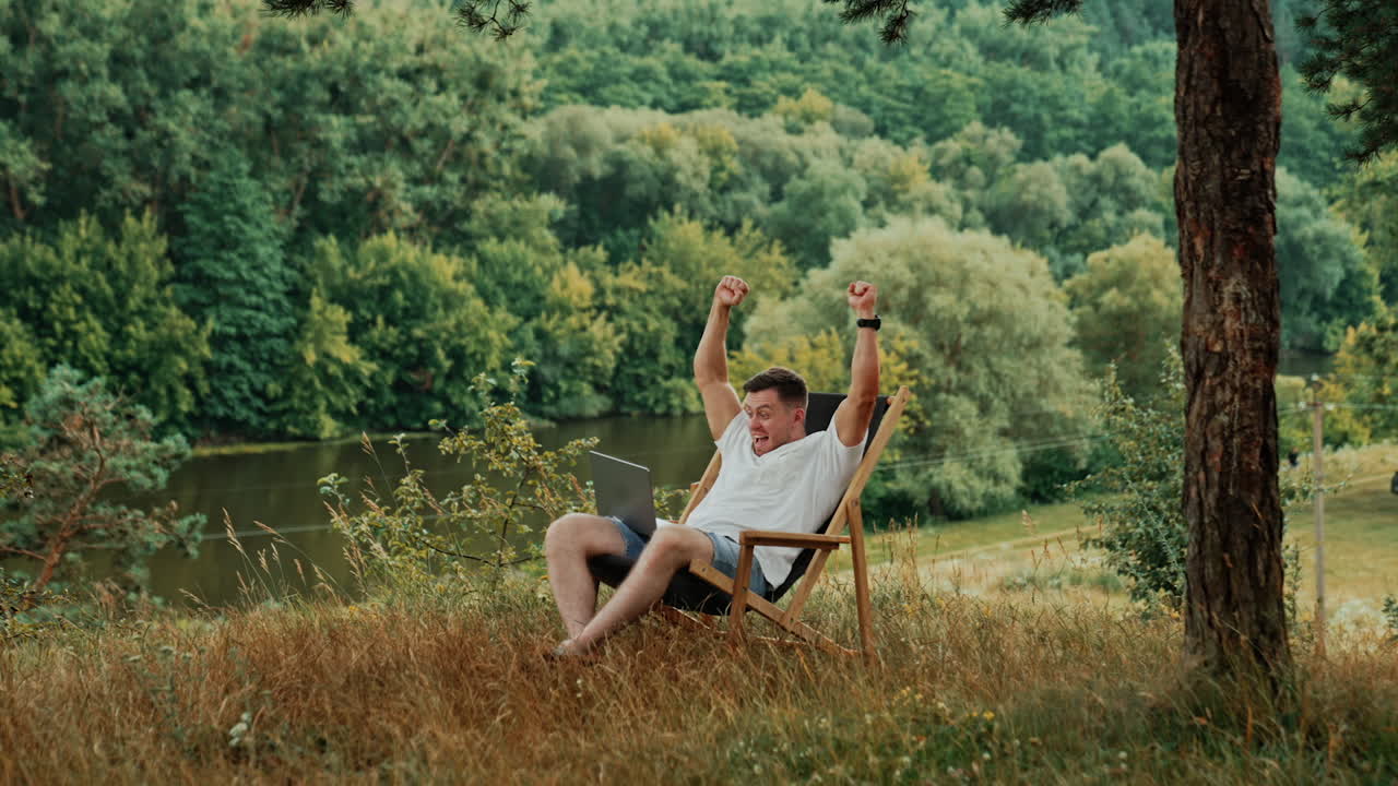 Caucasian man working remote in the beautiful nature in summer. Man looks at his laptop and raises hands cheering excitedly.