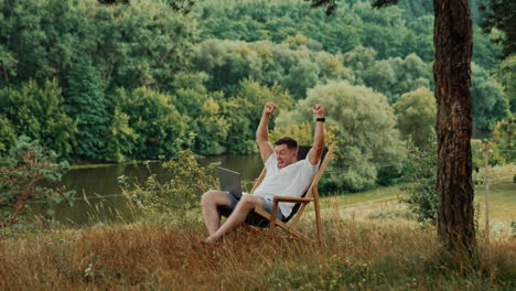 caucasian man working remote in the beautiful nature in summer. man looks at his laptop and raises hands cheering excitedly.
