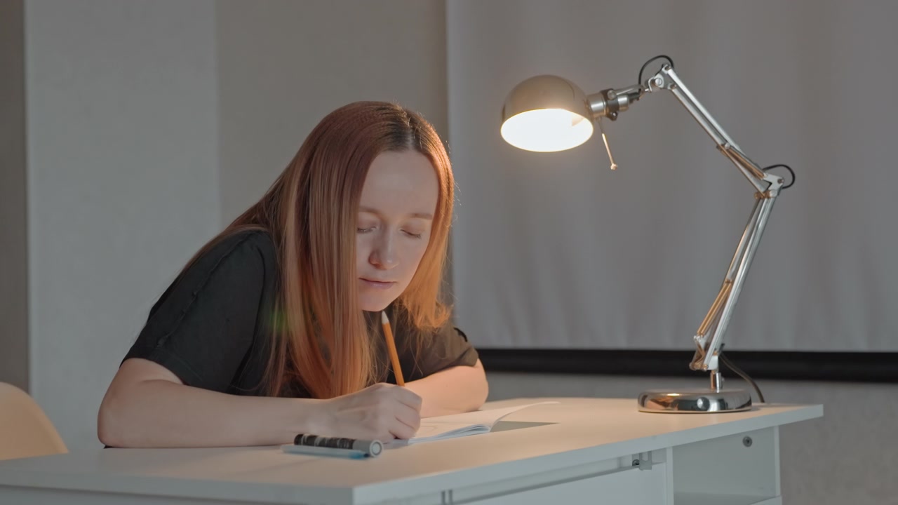 Young woman with long straight hair sits at white desk. Woman interacts with notebooks and pencils on table while maintaining focus. Background is plain with neutral colored walls