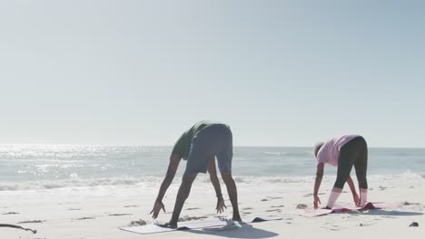 happy senior african american couple doing yoga and stretching at beach, in slow motion