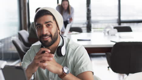 Un-Joven-Asiático-Con-Barba-Sonríe-A-La-Cámara,-Usando-Un-Gorro-Y-Auriculares-Alrededor-Del-Cuello,-