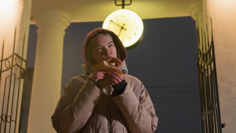 young woman in warm hooded jacket eating crispy snack outdoors under glowing clock at night, vintage architecture with illuminated clock contrasts with modern street scene