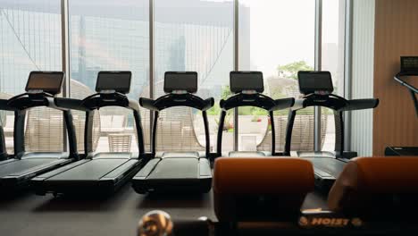 Panorámica-A-Través-De-Varias-Cintas-De-Correr-En-Un-Gimnasio,-Situado-Frente-A-Amplias-Ventanas-Que-Brindan-Una-Vista-Al-Exterior