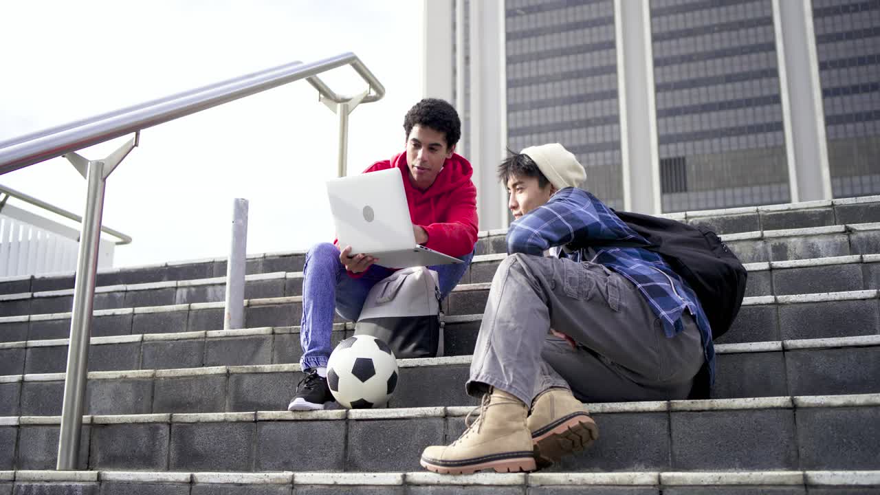 Diverse male friends on office steps tilting laptop toward friend pointing at screen reviewing info