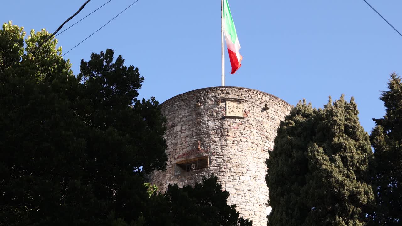 Bandiera tricolore italiana che sventola da un'antica torre di pietra a Bergamo Alta contro un cielo sereno