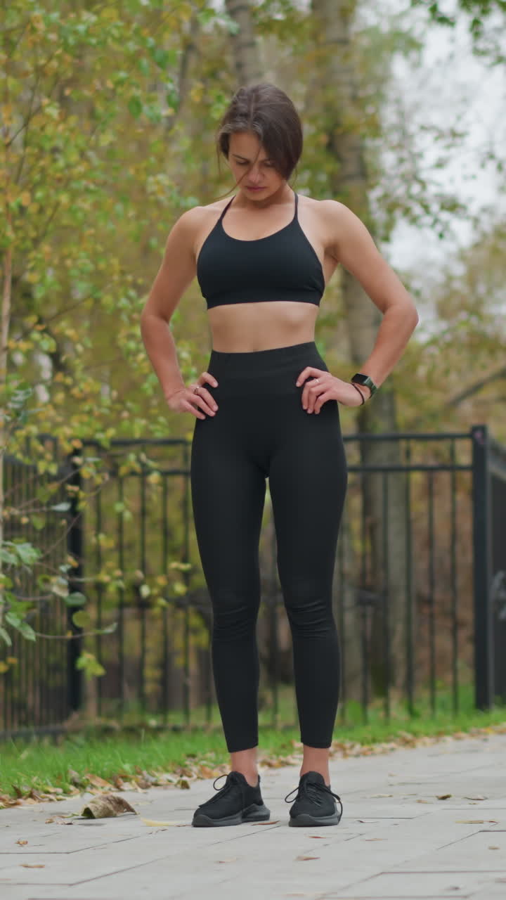 Lady in black neck top and leggings standing, stretching neck with hand on waist in park, blurred background featuring iron fence and trees, outdoor fitness, neck flexibility, posture correction