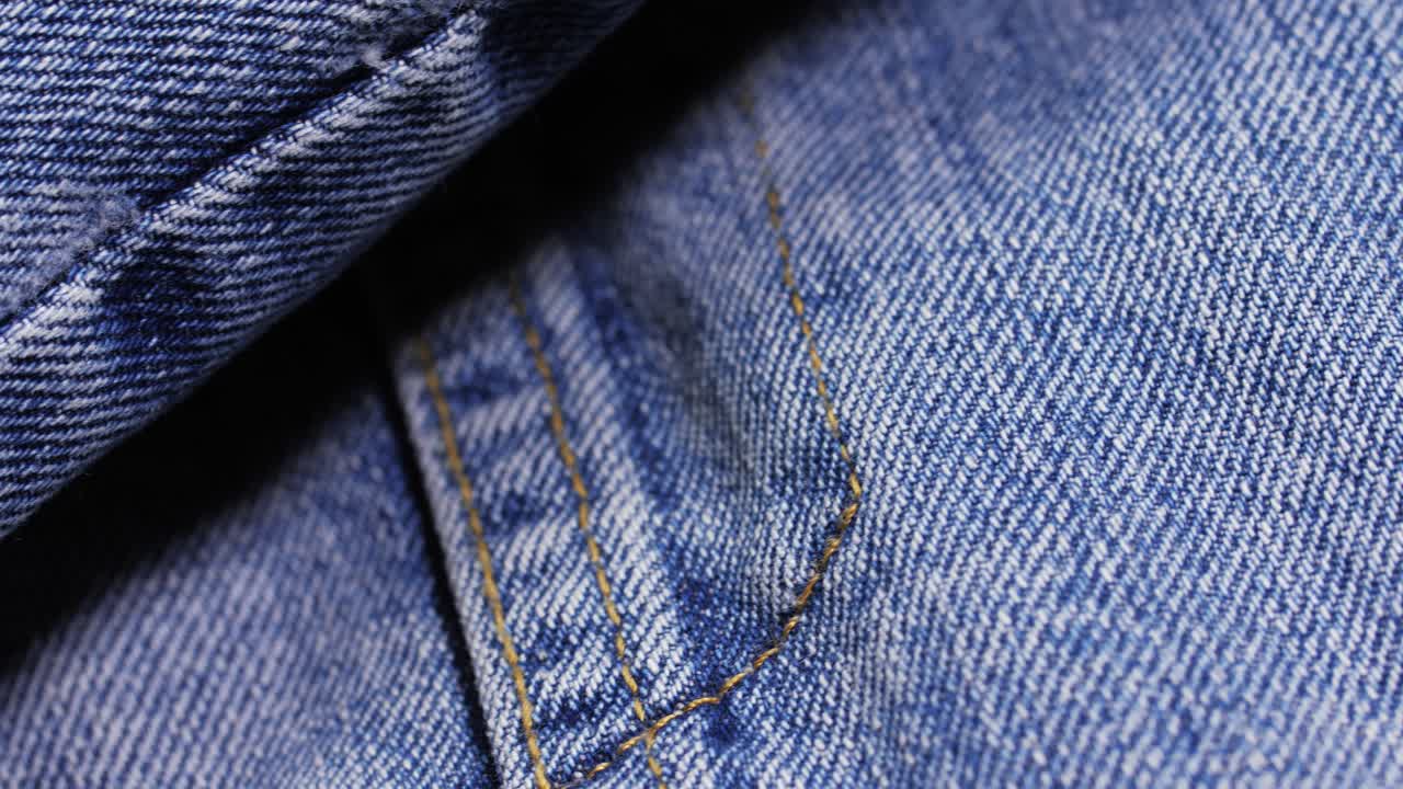 A blue denim jacket reveals twill texture, orange stitching and a metal rivet as the camera pans slowly left to right