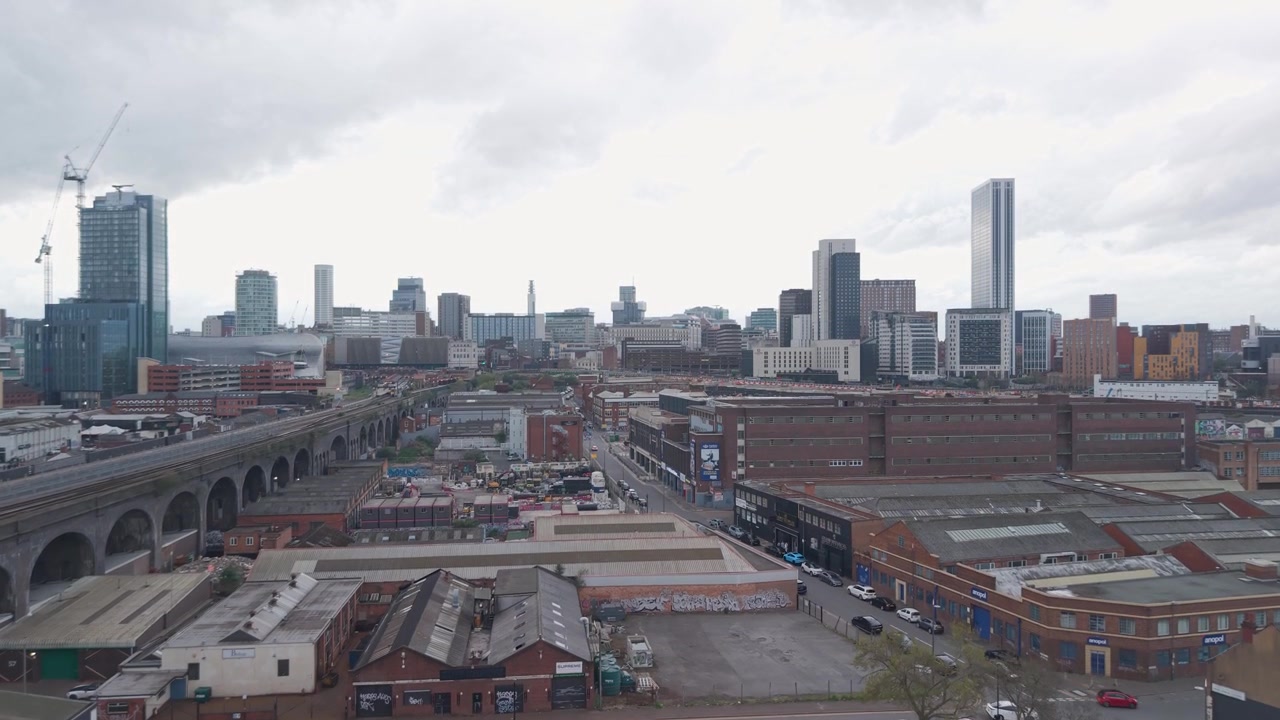 Skyline urbano con viadotti ferroviari, tetti industriali bassi e torri alte sotto un cielo nuvoloso