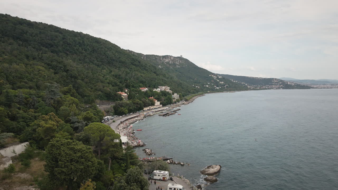 Drone cinematico 4K UHD scende sulla costa di Trieste sul Mare Adriatico, rivelando scogliere boscose, una costa curva e una strada costiera per promozioni di viaggio.