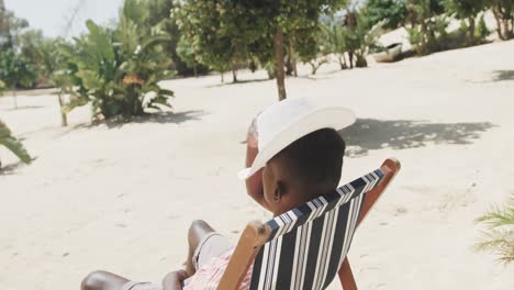 african american man sitting on deckchair, covering face with hat on sunny beach, slow motion