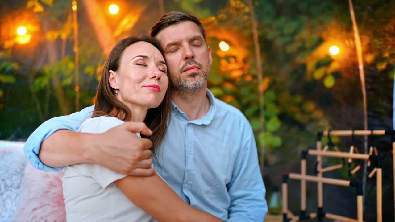 Um casal se abraçando de olhos fechados dentro de uma barraca de bolhas transparente em um acampamento de luxo. Floresta exuberante e iluminação no plano de fundo
