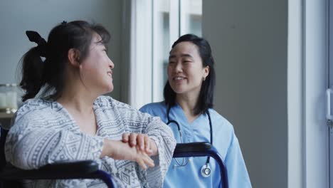 smiling asian female doctor looking out of window and talking with female patient in wheelchair