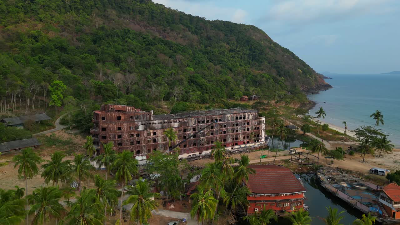 abandoned rusty ghost ship on Koh Chang on tropics island bay, Thailand, during sunset, with a crane working on it. Majestic aerial view flight wide orbit overview drone