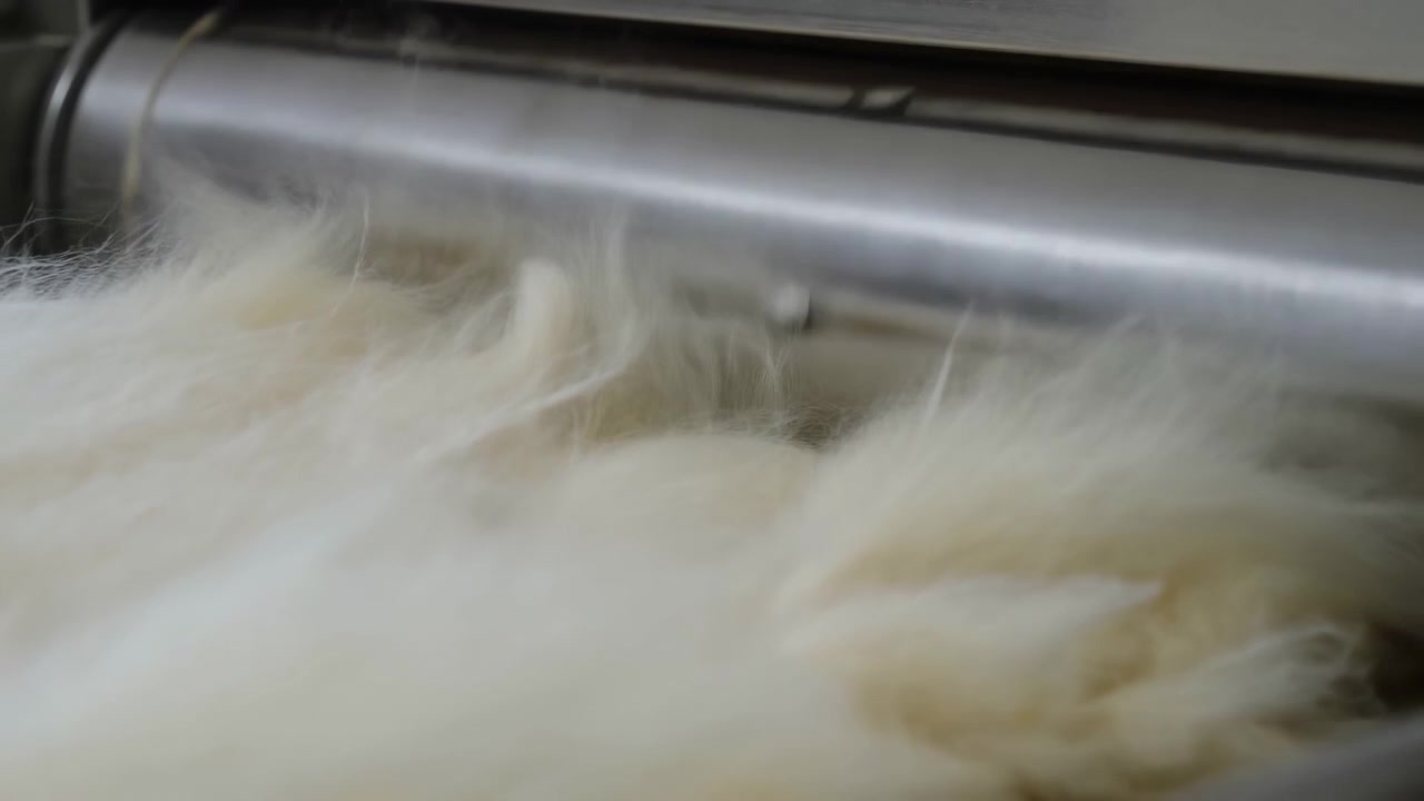 Primo piano di materiale bianco e soffice che passa sotto un rullo metallico in un ambiente di fabbrica, mostrando una consistenza morbida, movimento e lavorazione meccanica sotto una luce intensa