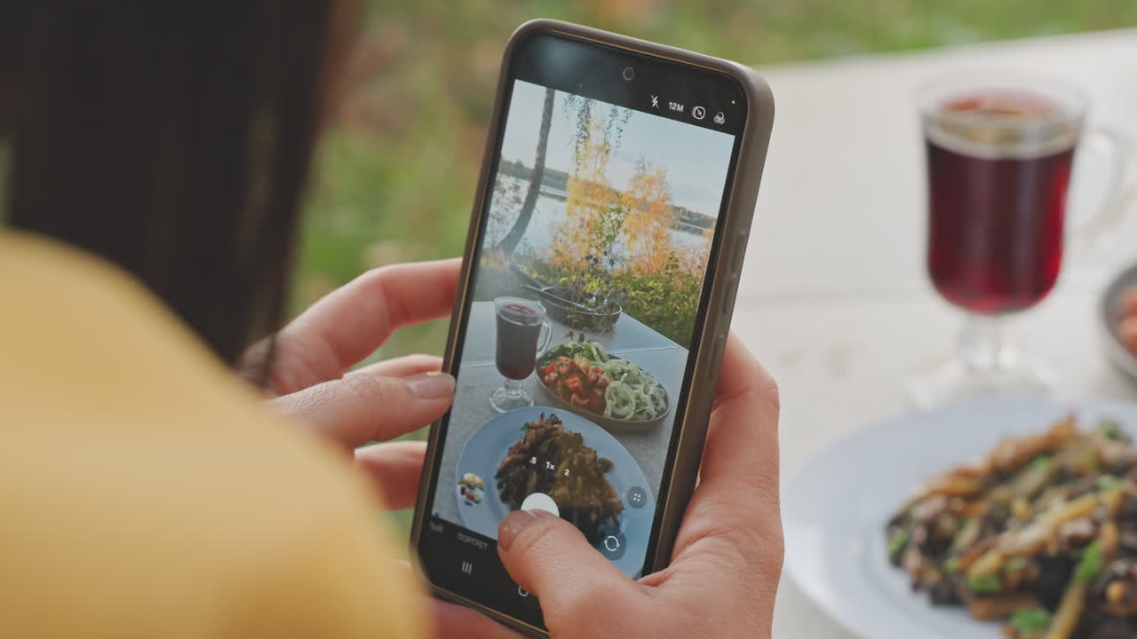 Phone closeup camping food photography showing woman hands framing plate and glass on portable table. Smartphone screen reflecting plated salad and drink, careful angle and filter preview visible