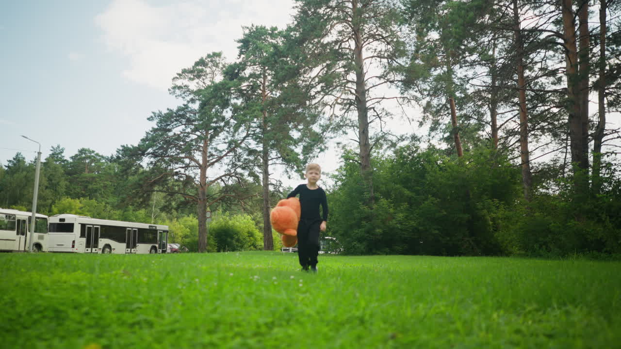 Little boy dressed in black holding oversized teddy bear while walking across grassy park, surrounded by lush trees with two white parked buses visible in the background under clear sky