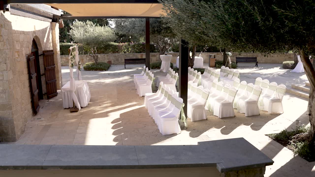 Le sedie sono disposte in file per gli ospiti a un matrimonio all'aperto. I tavoli sono apparecchiati nelle vicinanze, sotto una zona ombreggiata. L'ambiente è circondato da alberi e vegetazione, creando un bel sfondo.