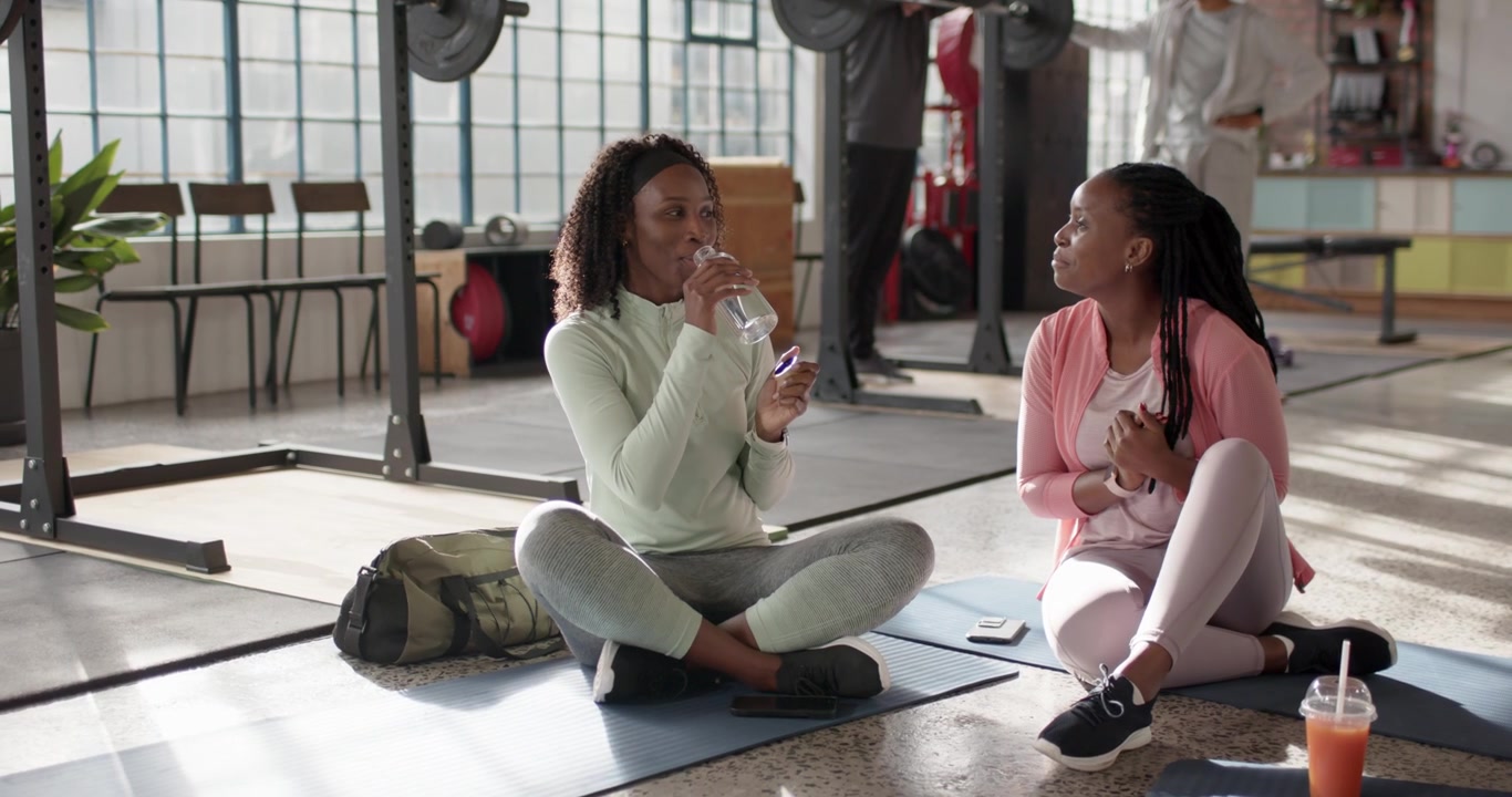 African American women in activewear resting between sets at gym, sipping water and checking phone
