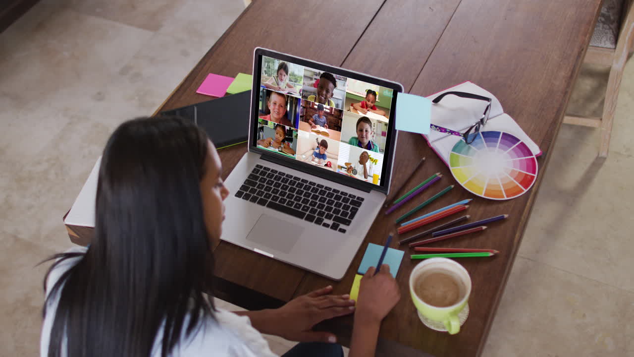 Maestra caucásica usando un portátil en videollamada con alumnos, tomando notas.