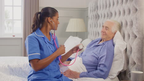 female care worker in uniform listening to chest of senior man at home in bed with stethoscope