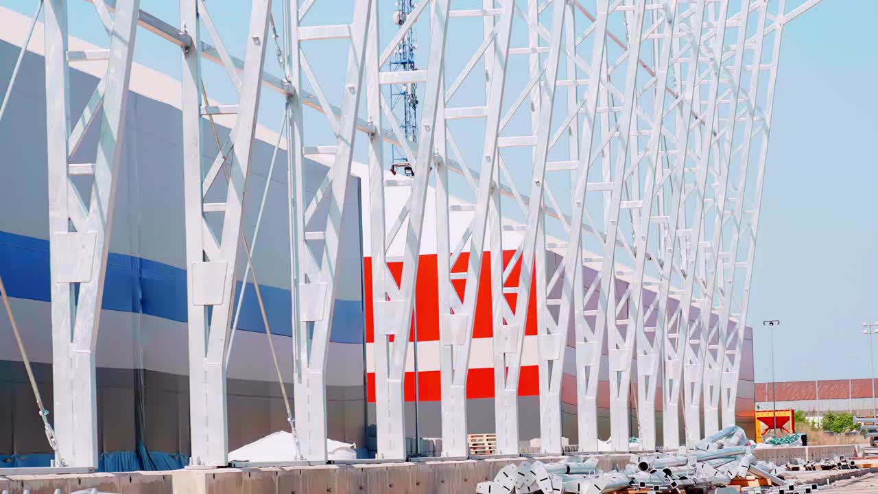 The construction site of a massive, modern warehouse or hangar featuring a striking white steel exoskeleton frame on a bright, sunny day, representing industrial growth and architecture