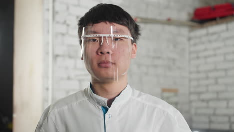 young lab scientist wearing lab coat and protective glasses nods head continuously in industrial setting, background features brick wall and red object